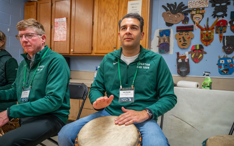 Juan Mundel wearing a Spartan Bus Tour long sleeve shirt sits and plays on an West African drum with artwork behind him on a blue wall.