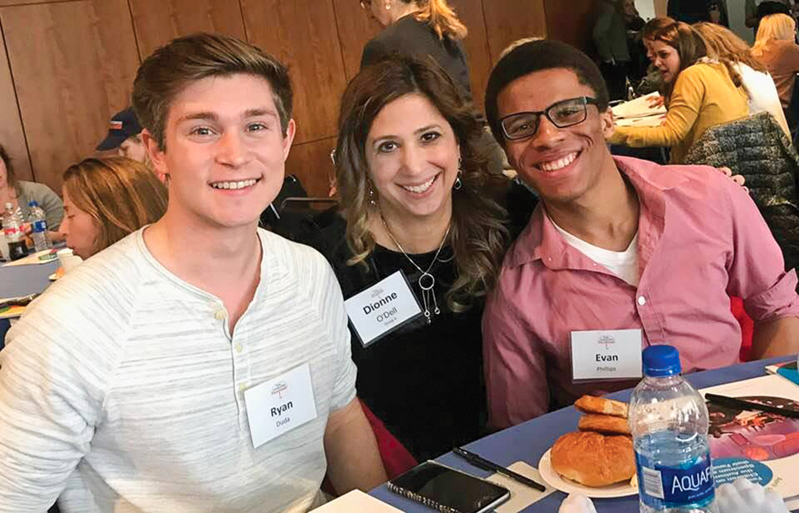 Three people sit smiling at a table during an event. They are wearing name tags that read "Ryan," "Dionne O'Dell," and "Evan."