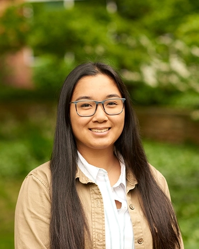 A headshot of a smiling person with long, dark hair and glasses. They are wearing a white striped shirt under a tan button-up jacket. The background is a soft-focus outdoor setting with green foliage and a hint of a brick building.