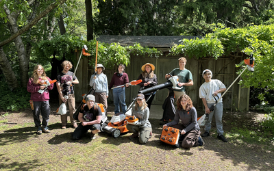 Group of 10 people pose outdoors in a garden courtyard with electric landscaping equipment, including string trimmers, leaf blowers, a lawn mower, hedge trimmers, and battery packs, in front of a wooden fence covered with green vines and surrounded by mature trees.