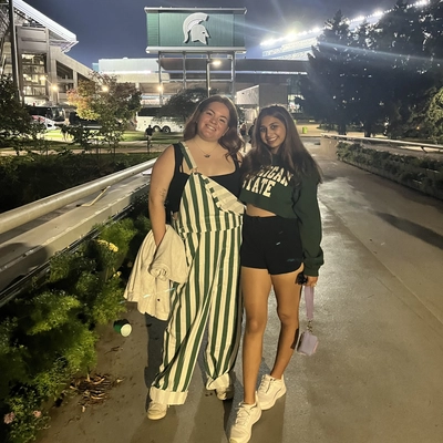 Two young women smiling in front of Michigan State University's Spartan Stadium at night.