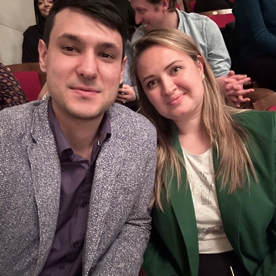 A photo of a man sitting in a gray jacket and a black shirt next to a woman in a green blazer and white shirt who has long brown hair.