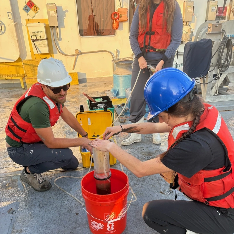 Researchers wear hard hats and life vests while filtering and collecting water samples into an orange bucket on a ship deck.