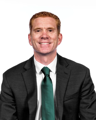 A man with short red hair wearing a black suit, white button down and dark green tie. He is sitting in front of a white background.