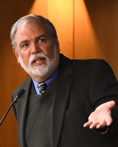 Sherman Garnett speaks into a microphone, gesturing with an open hand in front of a wood-paneled wall.