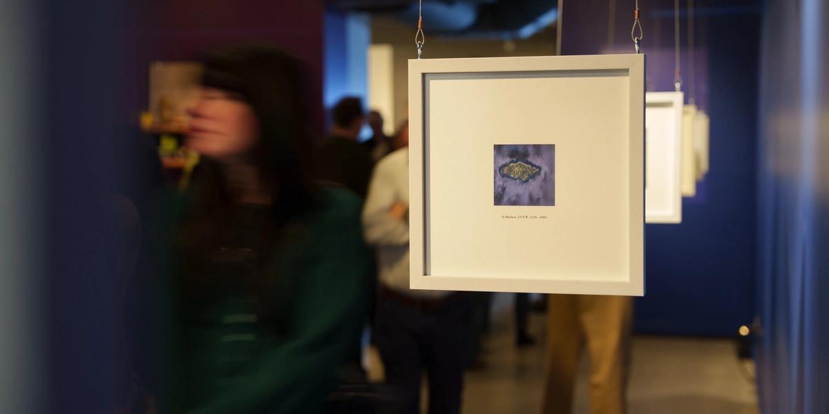 A framed artwork hangs from the ceiling on thin cables in a gallery space, showing a small abstract image centered on a white mat. More framed pieces hang in a line behind it. Visitors move through the gallery in the background, with colorful walls and soft lighting creating a lively exhibition atmosphere.