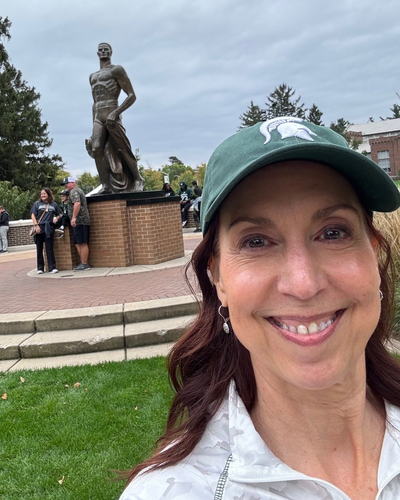 Person wearing a green Spartan helmet cap takes a selfie on campus with the Spartan statue.