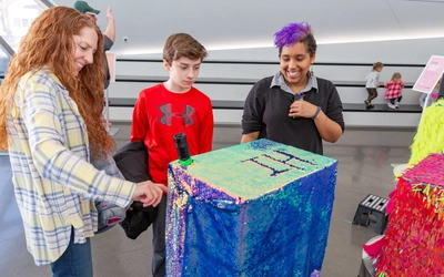 Three students engage with an interactive sequin sculpture display at museum.