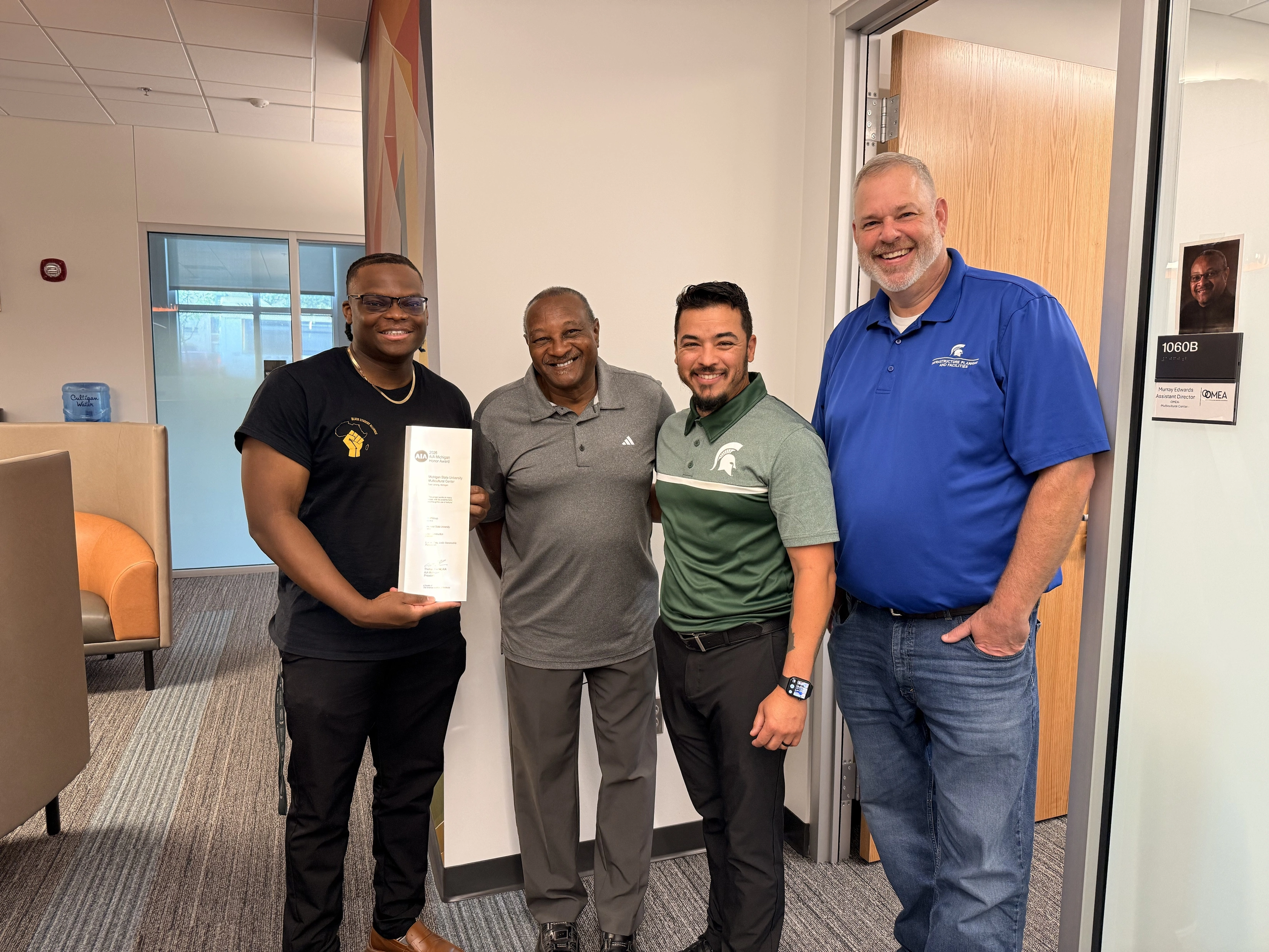 a group of men, staff employees standing holding up a framed award letter. AIA Michigan Building Award, MSU, Multicultural Center