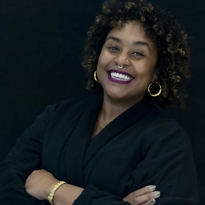 A portrait of a Black woman with curly, highlighted hair smiling warmly with her arms crossed against a solid black background. She is wearing a black wrap-style top, large gold hoop earrings, a gold bracelet, a septum ring, and bold purple lipstick.