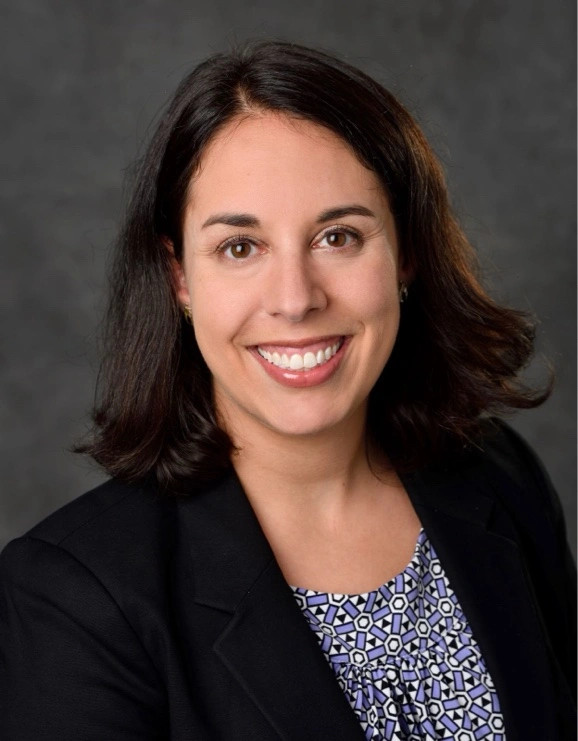 A woman with shoulder length black hair, brown eyes, and light skin smiling and wearing a black jacket over a mosaic print purple and white top.