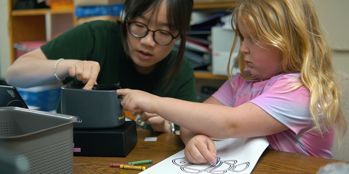 A researcher and a elementary student work at a desk with crayons and papper