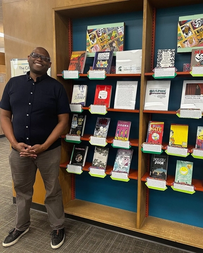 Julian Chambliss at the Capital Area District Libraries downtown Lansing location next to the display of books created by his Readings in Graphic Narratives class.
