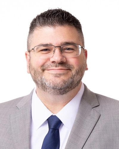 A man wearing a light patterned gray blazer with white button down and blue tie against a white background. He has a brown beard and glasses.