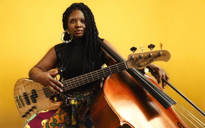 Endea Owens poses with a guitar and stand up bass in front of a yellow background.