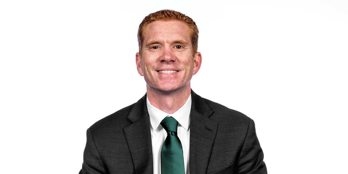 A man with short red hair wearing a black suit, white button down and dark green tie. He is sitting in front of a white background. 