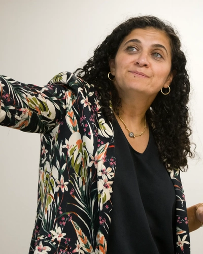 Linda Sayed, wearing a floral jacket over a black top, gestures with one arm extended toward the left side of the image.