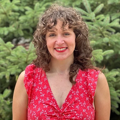 A woman with curly brown hair smiles at the camera while standing outdoors in front of green trees, wearing a red sleeveless dress with a small white pattern.