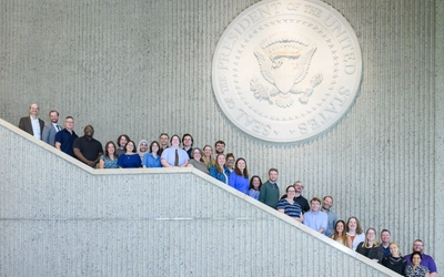 A large group of people poses on a grand indoor staircase beneath a prominent, large carving of the Seal of the President of the United States on a textured wall.