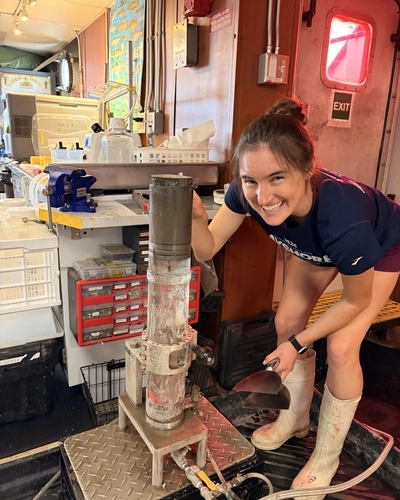 Paige Kohler, PhD student, smiles in a shipboard laboratory setting while handling sampling equipment next to a freshly extracted sediment core tube.