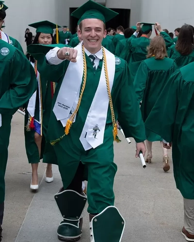 Graduating student in a green gown wearing Sparty mascot boots while walking in a commencement procession.