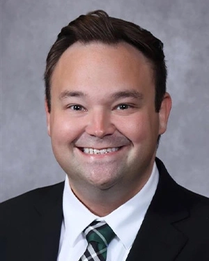 Professional headshot of a smiling man with short dark brown hair, wearing a black suit jacket, a white collared dress shirt, and a green, white, and black patterned tie against a plain gray studio background.