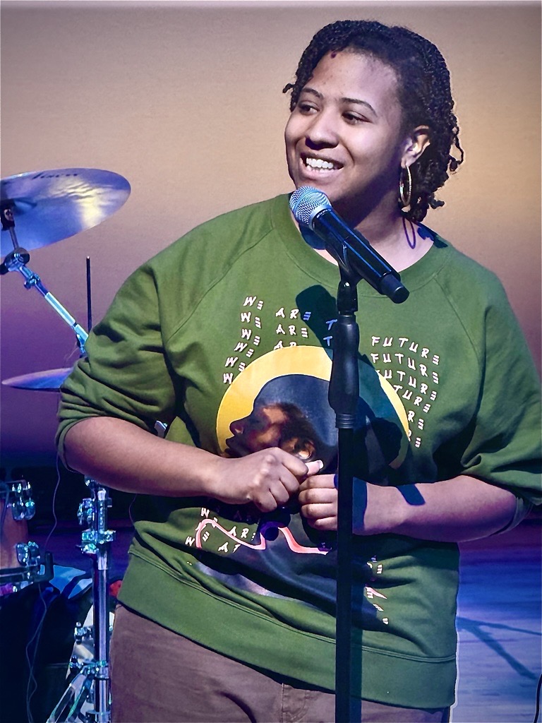 A woman stands in front of a mic on stage with a drum kit in the background
