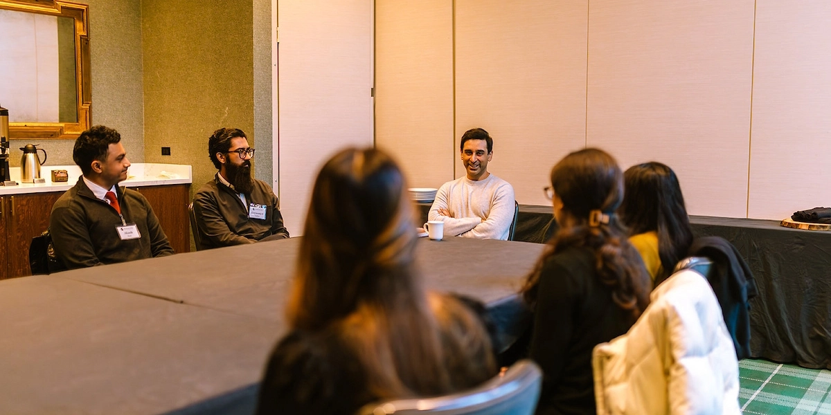 Six people sit around a conference table, with Dhaval Bhanusali in a white shirt, arms crossed smiling at the head of the table
