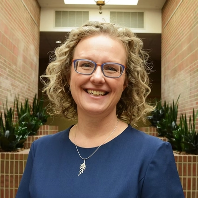 A smiling woman with curly blonde hair and blue-framed glasses, wearing a navy blue top and a silver leaf-shaped necklace, standing in a brick-walled hallway with potted plants in the background.