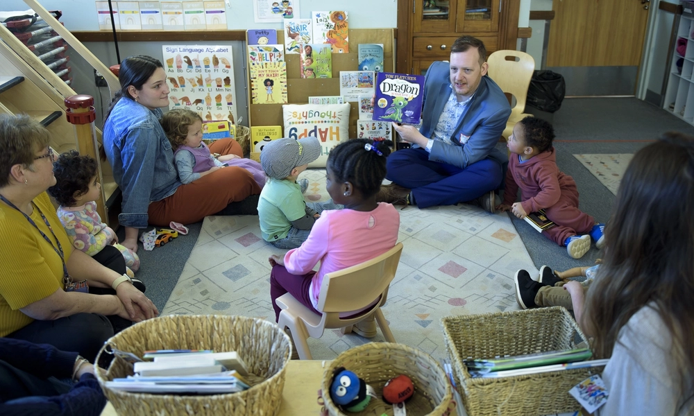 Man sits on the ground surrounded by a circle of children reading them a book.