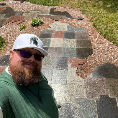 A bearded man smiling in a white Spartan hat. Behind him is a large stone map of Michigan on the ground.