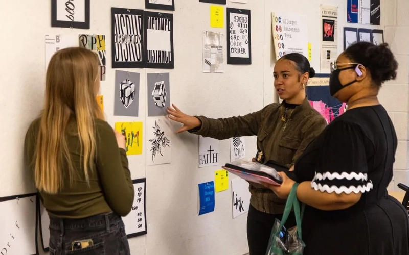 Three students discuss graphic design artwork, some black and white and others yellow or blue artwork, pinned to a white gallery wall.