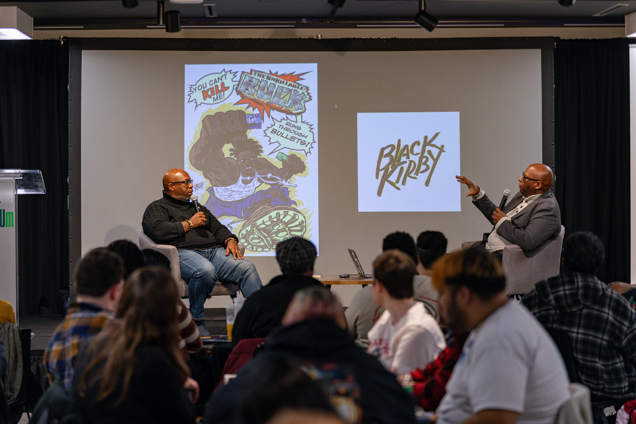two panelists in discussion in front of a screen with Black Kirby comic projections in the background and students in the foreground