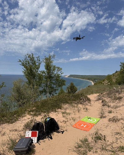 Equipment with backpacks on the ground and drones in the air on a sand dune above Lake Michigan.
