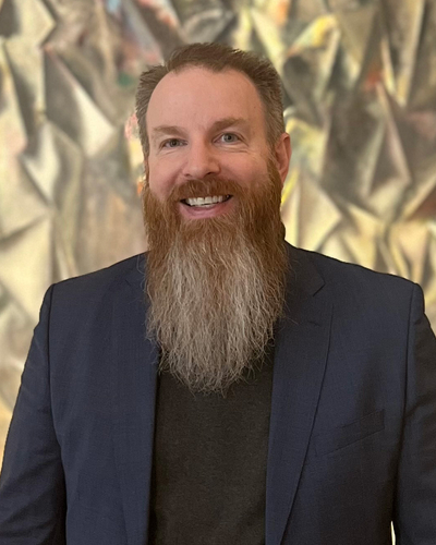 A smiling man with a long brown and gray beard wears a dark blazer over a black shirt while standing in front of an abstract, softly lit background.