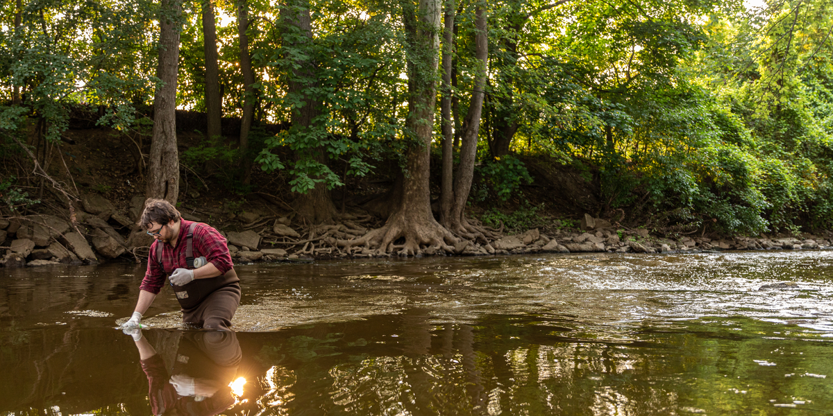 A person stands knee-deep in a river, collecting water samples with a container. Surrounding environment includes dense green trees and a rocky riverbank.