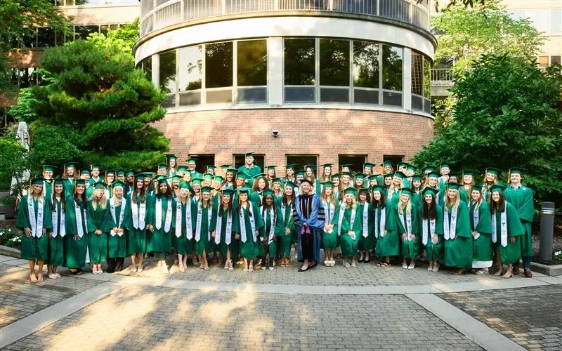 A large group photo of nursing students gathered outdoors on a paved courtyard for the Michigan State University College of Nursing Pinning Ceremony. The students are dressed in bright green academic gowns, mortarboard caps with tassels, and white stoles draped around their shoulders, with a faculty leader standing centrally in dark academic regalia and a doctoral hood. They are positioned in rows in front of a curved brick campus building surrounded by lush green trees, shrubs, and potted flowers under soft daylight.
