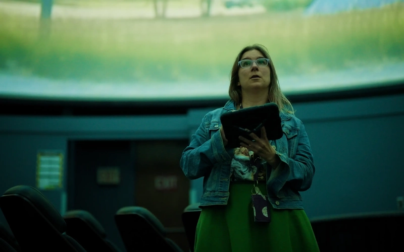Shannon Schmoll stands in a planetarium holding a tablet, looking up at a projected field.