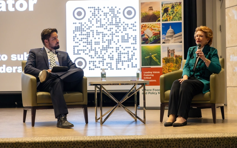 Two speakers sit on armchairs on a stage beside a QR code presentation screen at a lecture event.