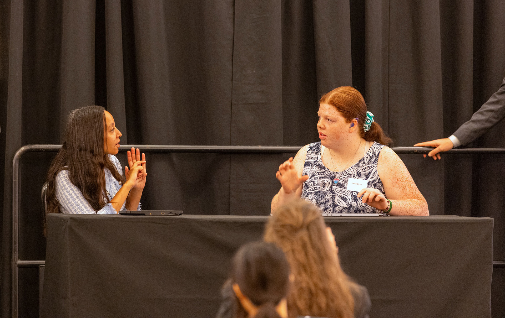 Two women sit at a table on stage, having a discussion using hearing assistive devices with audience members in the foreground