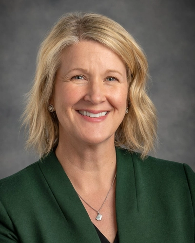 A woman with blonde hair, wearing a green blazer and black shirt underneath and a silver necklace sitting in front of a gray background.