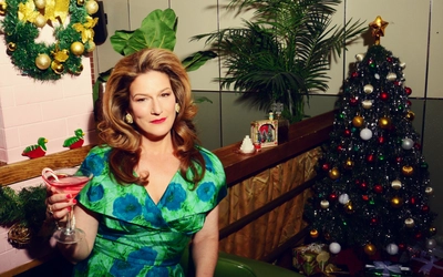 Ana Gasteyer poses with a martini in hand. A Christmas tree and palm tree with a wreath on a wall are seen in the background.
