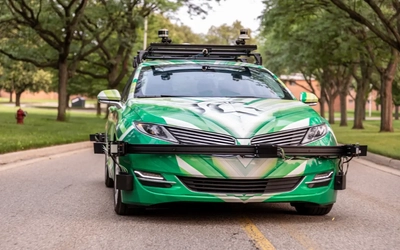Autonomous research vehicle with LiDAR and camera sensors driving on a campus road.