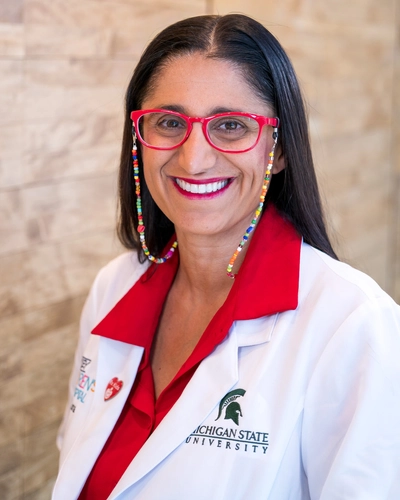 Dr. Mona Hanna-Attisha smiling in a white Michigan State University lab coat and red-rimmed glasses.