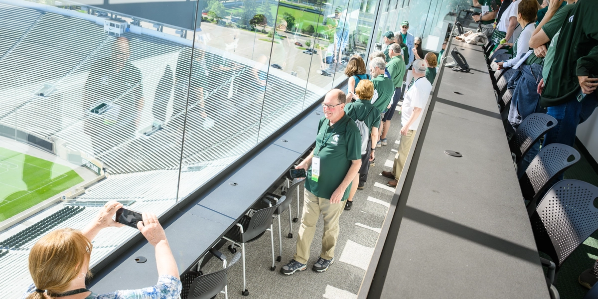 Visitors wearing Michigan State gear stand inside a suite overlooking the field at Spartan Stadium. Several people are taking photos with their phones through the large glass windows.