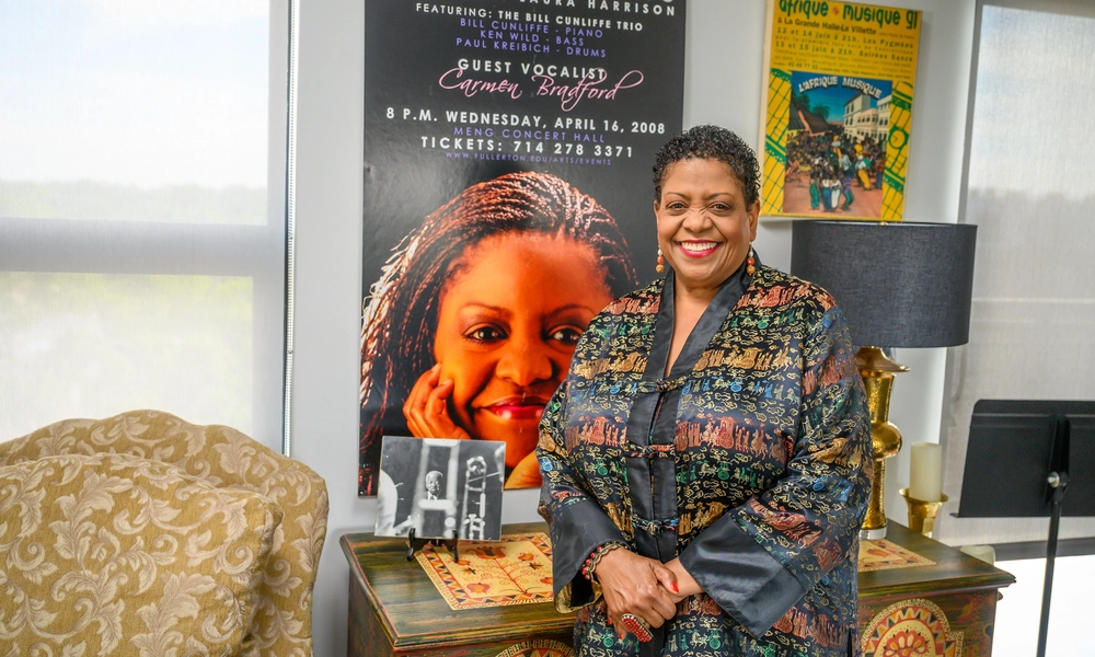 Person standing beside a decorative cabinet in front of a large concert poster featuring a close-up portrait and the text “Jazz Singers” and “Guest Vocalist Carmen Bradford.” A framed poster, table lamp and small black-and-white photograph are visible in the room.