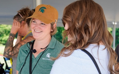 Caitlin Splawski wearing an MSU Extension polo shirt and hat smiling at an event participant.