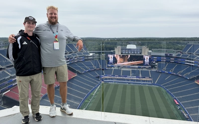 Two field crew members stand beside a glass railing overlooking Gillette Stadium’s soccer pitch and empty stands.