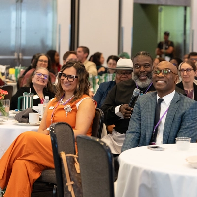 A crowded conference hall filled with people sitting around round banquet tables with white tablecloths. In the foreground, a woman in a bright orange jumpsuit and a man in a blue patterned suit jacket are seated facing forward, smiling during a presentation. Behind them, another man in a black tunic holds a microphone, smiling toward the front of the room.