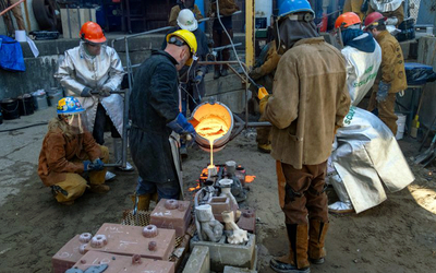 People in protective jackets, gloves, and helmets work together pour molten metal into molds in an industrial setting outside.